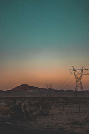 A vertical  shot of transmission towers connected by wires in the desert with mountains in the backgroundの写真素材