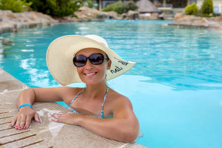 A tanned female wearing a straw hat and sunglasses smiling in a pool