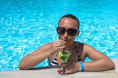 A young female in a pool drinking a glass of green delicious cocktail under the sunlight