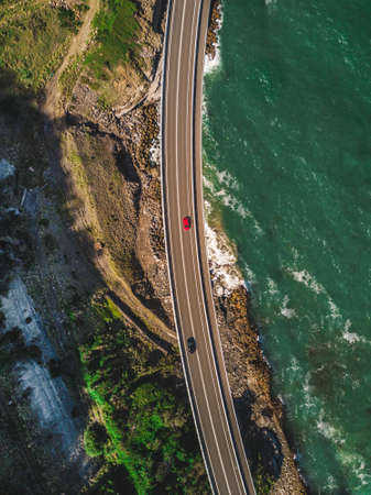 A narrow curvy road with cars alongside green mountains built at the coast of the seaの写真素材