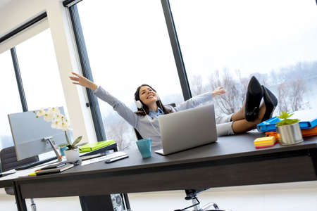A businesswoman listening to music happily with headphones with legs on the table during the break