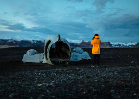 A photographer in a yellow raincoat taking a picture of an old broken airplane part in the evening with cloudy blue skies in the backgroundの写真素材