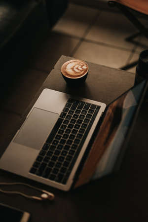 A high angle shot of a laptop with a beautifully made cappuccino with flower art on the side