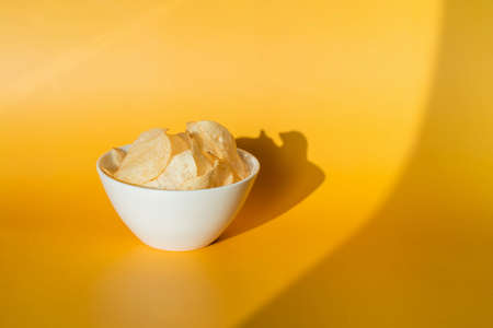 A closeup shot of plain potato chips in a white glass bowl on a yellow background