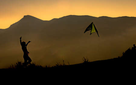 The silhouette of a kid flying a kite surrounded by mountains during the sunsetの写真素材