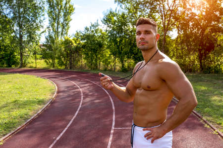 A shirtless fit man using a stopwatch on a running track in the daytime