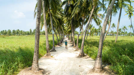 A young man standing in the middle of a sandy road with palm trees on both sides on an island in...