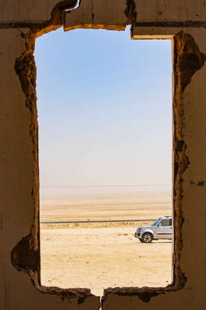 A vertical shot of a square hole on a wall with a view of a car parked in the desert with clear sky in the background at daytimeの写真素材