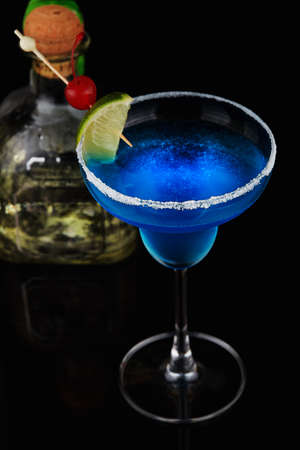 Exotic blue cocktail with lime and cherry in a classic cup with bottle with glass reflection on black backgroundの写真素材
