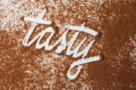 A closeup of [tasty] written with brown powder on a white surface under the lights