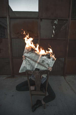 A vertical shot of a male sitting on a chair reading a burning newspaper 
Concept- Fake news
