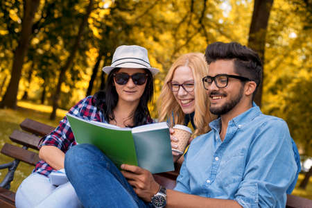 A group of young friends reading a book and smiling on a park bench
