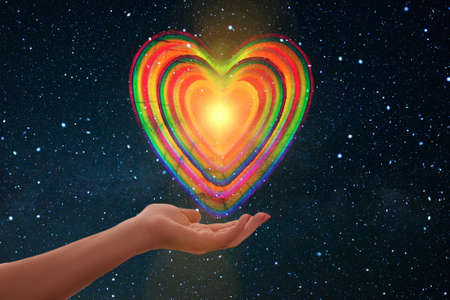 A closeup shot of a female hand holding a colorful heart with cosmic backgroundの写真素材