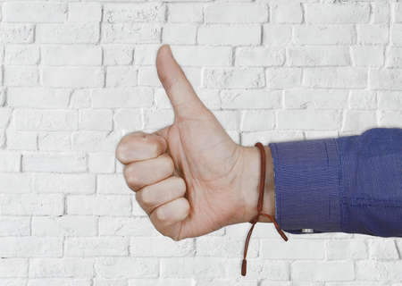 A closeup of a person doing the thumbs-up gesture with a white wall on the background