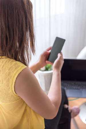 A vertical shot of a woman sitting on a chair holding her phone with a laptop  on a table near the...