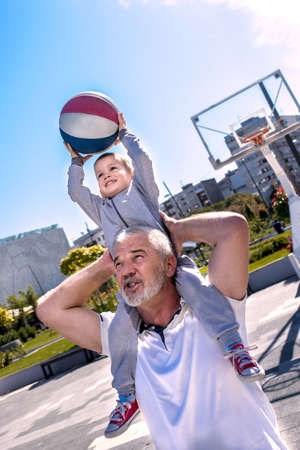 A vertical shot of a grandson sitting on his grandfather's shoulders playing basketball