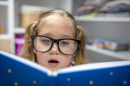 An adorable little girl with glasses reading a book with a surprised facial expression