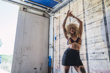 An attractive young Caucasian female with blonde curly hair and tattoos wearing a crop top and a...