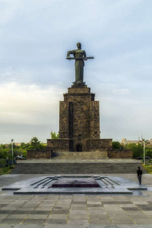 Old statue of Mother Armenia holding a sword on a tall stone pedestal with a fountain and a person standing in front of itのeditorial素材