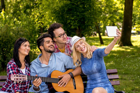 A group of young people taking a selfie and playing the guitar  while hanging out in the park