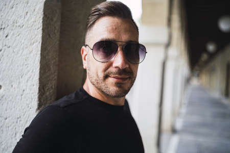 A young Caucasian male with sunglasses wearing a black shirt and jeans standing in a hall with...
