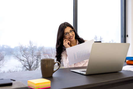 A young businesswoman talking on her phone and holding a business document while  working in her...