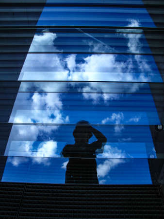 A vertical shot of glass facades with the reflection of person holding camera under clear blue skyの写真素材
