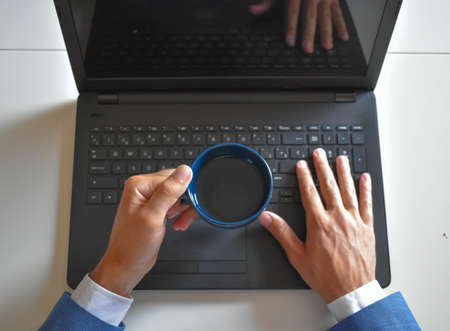 An overhead shot of a businessman in formal attire holding a mug and typing on a computer keyboardの写真素材