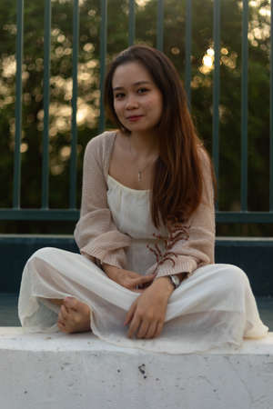 An attractive Asian young female with long hair wearing a white dress with a jacket sitting near a metal fenceの写真素材
