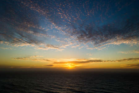 The beautiful wavy ocean gleaming under the sunset piercing through the cloudsの写真素材