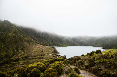 An aerial shot of a pond surrounded by green hills and forested mountain in a fog