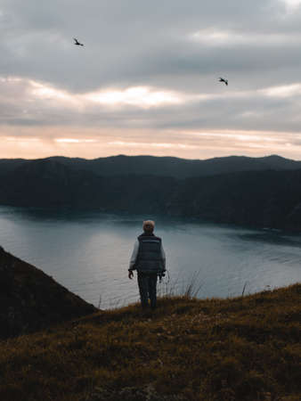 A vertical shot of a person standing on grass by the water near mountains under a pinkish sky with...