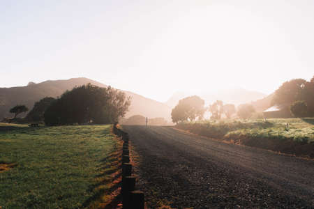 A narrow countryside gravel road in the middle of a green field with trees and hills in the backgroundの写真素材