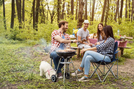 A shallow focus shot of four young caucasian friends having a barbecue picnic in a forest with a dogの写真素材