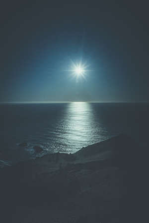 A vertical shot of a beautiful reflecting sea and the shining moon in the starry skyの写真素材