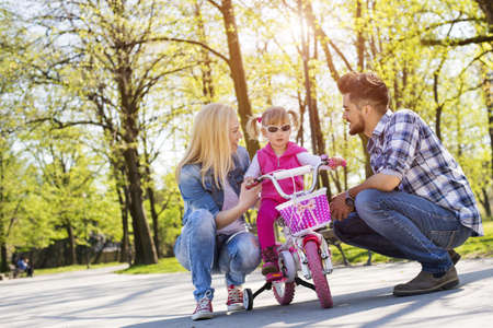 A young couple teaching their adorable daughter to ride a bike in a park on a sunny dayの写真素材