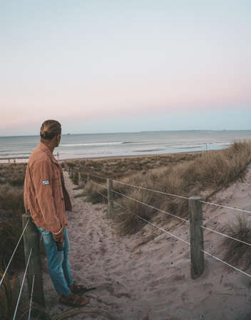 A vertical shot of a male leaning against a fence on the beach with a sea and a clear sky in the...