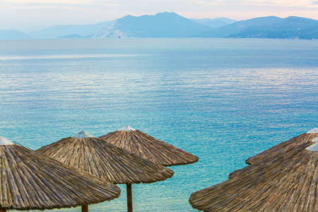 A wide shot of wooden beach umbrellas near clear ocean surrounded with mountains in the distanceの写真素材