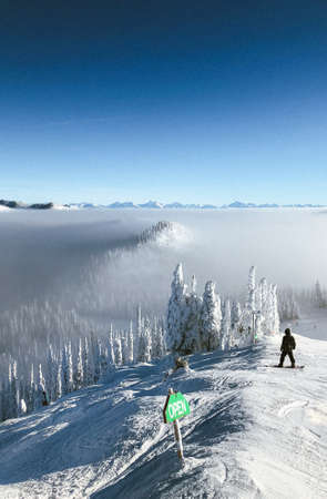 A vertical shot of a person with a snowboard on a snowy mountain above the clouds with a clear sky in the backgroundの写真素材