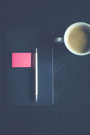 A vertical overhead shot of a gray click pen on a gray notebook near a cup of coffee on a black...
