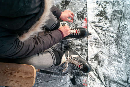 YOUNG MAN SEATED IN A WOODEN CHAIR WITH A FUR COAT LACING ICE SKATES IN A OUTDOOR BY THE ICE OF...