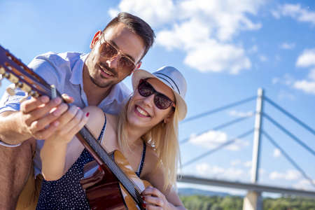 A shallow focus shot of a Caucasian couple playing a guitar under blue sky and white clouds