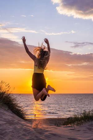 A vertical shot of a female jumping on a beach shore with a beautiful sky in the background