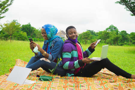 A happy Nigerian couple using their laptops and cellphones while on a picnic on a grassy meadow