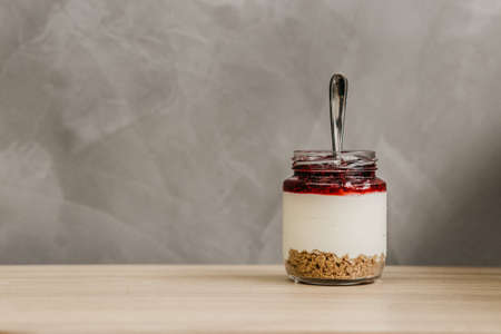 A closeup shot of a glass jar full of cereal, yogurt, and fruit jam with a spoon inside
