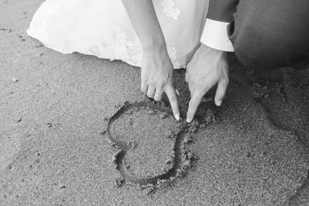 A grayscale closeup shot of a couple showing love for one another by drawing a heart on the sandの写真素材