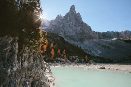 A wide shot of high rocky mountains and brown-leafed trees near the water with sun raysの写真素材