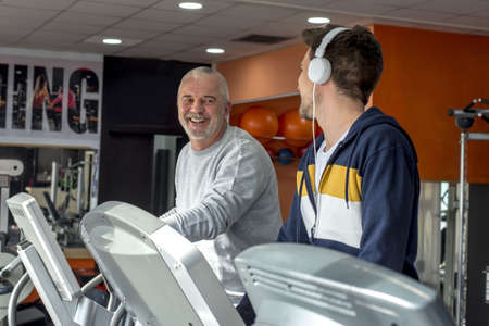 A young male in headphones and a senior male exercising together on the treadmills at the gym