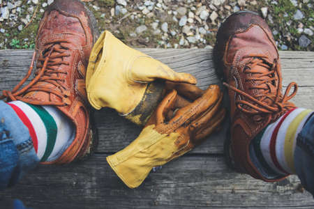 A horizontal shot of human feet with brown shoes and a pair of yellow gloves on a wooden benchの写真素材