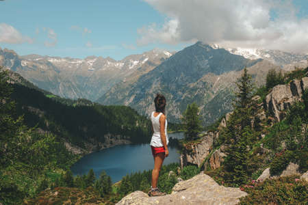 A wide shot of a female standing on a rock on top of a mountain facing the beautiful view of the lake and the mountains covered in snowの写真素材
